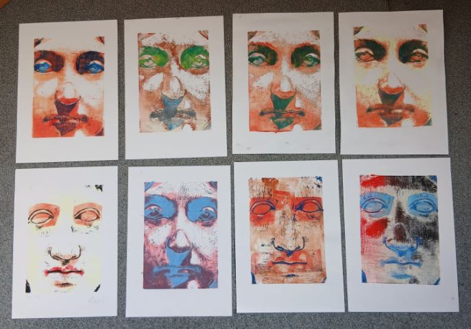 A view of eight finished prints. The ancient face appears in many different colours, ranging from pale yellow through orange, blue and green to dark brown.