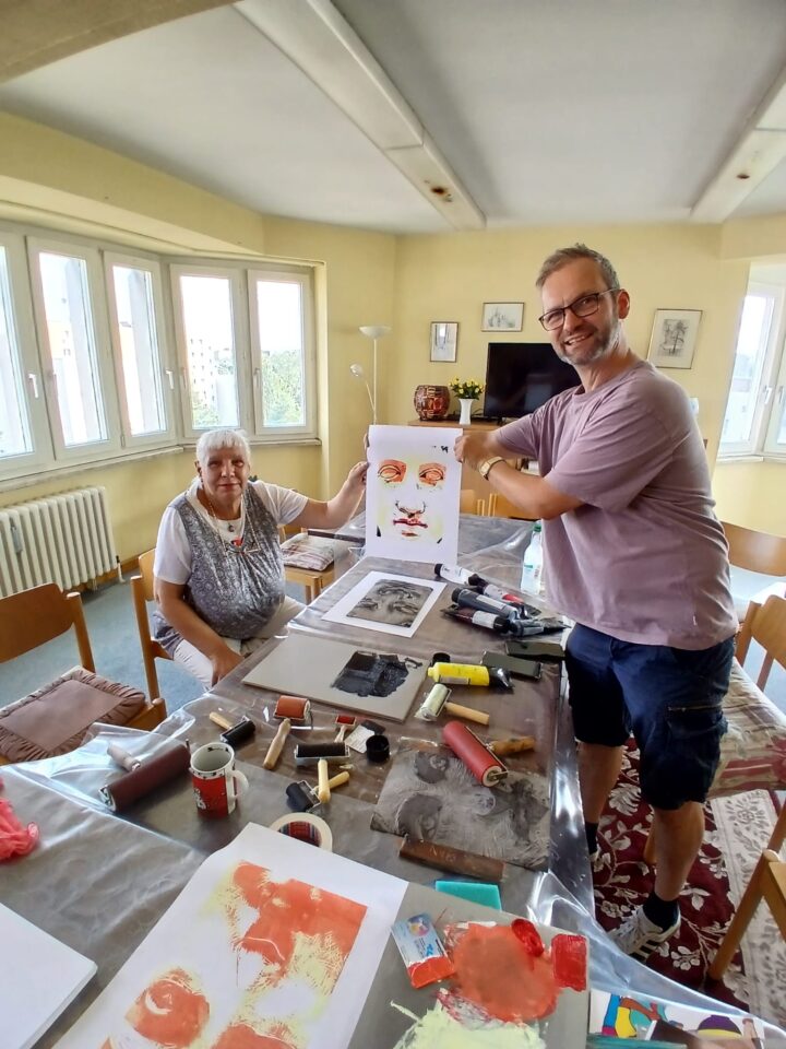 A man wearing glasses and an elderly woman are holding up a printed picture together, featuring a colourful, antique-style face, and smiling at the camera.