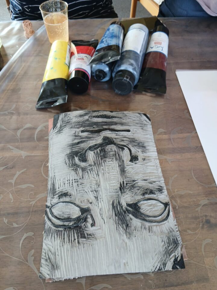 A view of the printing materials on the table. Tubes of paint in yellow, red, blue, white and brown. A linocut plate with an antique face carved into it.