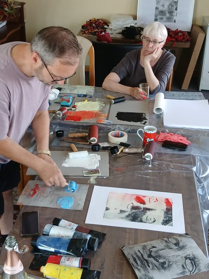 A man wearing glasses is rolling a light blue paint roller over a grey linoleum sheet. An ancient face is carved into the sheet. The first print is already lying at the right-hand edge of the picture. An elderly woman is watching the man.