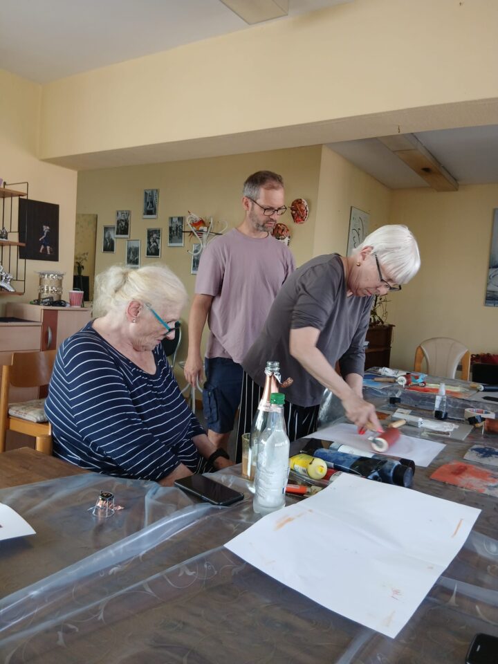 An elderly woman is standing at a table, rolling a sheet of paper with a paint roller. A man wearing glasses – the artist – and another elderly woman are watching her.