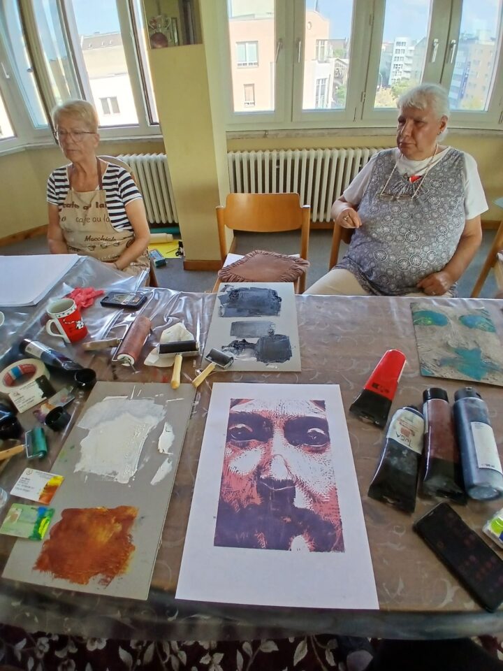 A view of the workbench. The first print can be seen, alongside materials such as inks and rollers. Two elderly women are sitting at the table on the opposite side.
